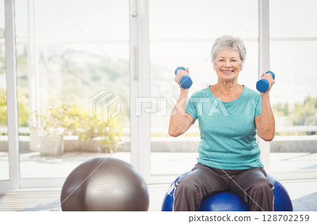 Smiling senior woman lifting blue dumbbells in home gym, with stability ball and glass windows Smiling senior woman lifting blue dumbbells in home gym, with stability ball and glass windows 128702259