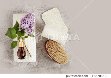 Spa treatment flat lay with bottle of organic oil essence collagen. Dropper bottle with natural serum on a white towel with lilac flowers, massage brush and loofahs on grey background. Copy space 128703389