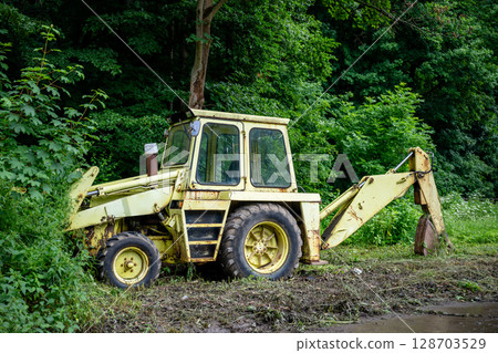 Old yellow backhoe loader parked at forest edge with overgrown vegetation Old yellow backhoe loader parked at forest edge with overgrown vegetation 128703529