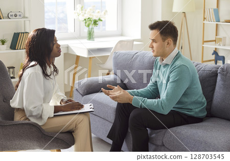 Male client sitting on sofa, talking to female psychotherapist during professional therapy session. 128703545