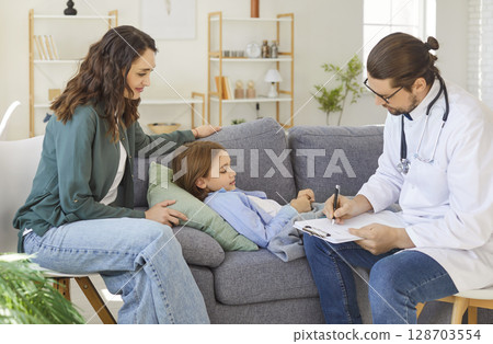 Male doctor writing prescription during home visit while child resting on sofa with mother nearby. Male doctor writing prescription during home visit while child resting on sofa with mother nearby. 128703554