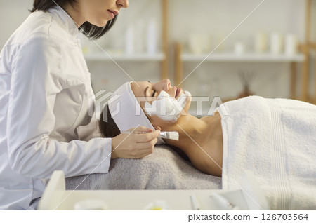 Professional cosmetologist applying moisturizing mask to young woman lying on couch in spa salon. 128703564