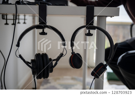 Two pairs of headphones hanging on desk-mounted holders in modern home office workspace 128703923