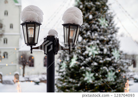 Snowy lantern with decorated Christmas tree and church in blurred background, shallow DOF Snowy lantern with decorated Christmas tree and church in blurred background, shallow DOF 128704093