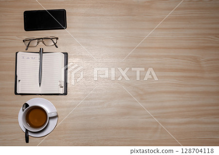 Desk with eyeglasses, notepad, smartphone, pen and a cup of tea on a wooden table. Top view with copy space. Flat lay - image 128704118