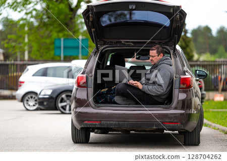 a man sitting in the open trunk of a car and working with a laptop, mobile technology, remote work 128704262