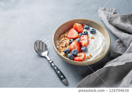 Natural yogurt with granola, blueberry and strawberry in a bowl on a blue background with spoon  128704638