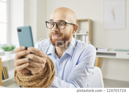 Obsessed man employee sitting in office with his hands tied with rope addicted to mobile phone Obsessed man employee sitting in office with his hands tied with rope addicted to mobile phone 128705728