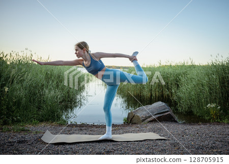 Young woman doing yoga exercise outdoors near lake in summer  128705915