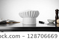 A pristine chef's hat rests on a stainless steel surface, accompanied by culinary tools and a cookbook. 128706669