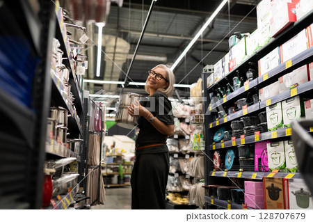 Stylish mature woman shopping for home essentials in a modern retail store selecting cookware and Stylish mature woman shopping for home essentials in a modern retail store selecting cookware and 128707679