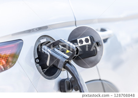 Focus closeup electric vehicle plugged in with EV charger device from blurred background of public charging station powered by renewable clean energy for progressive eco-friendly car concept Focus closeup electric vehicle plugged in with EV charger device from blurred background of public charging station powered by renewable clean energy for progressive eco-friendly car concept 128708848
