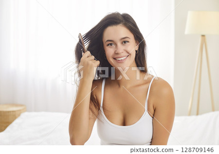 Young woman brushing hair sitting on bed in bedroom after waking up in morning Young woman brushing hair sitting on bed in bedroom after waking up in morning 128709146