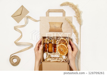 Female hands hold cardboard gift box with self care set with natural cosmetic on paper bag. Packaging goods from online store and delivery. Eco friendly packaging concept. Ecologic gift set. Flat lay 128709261