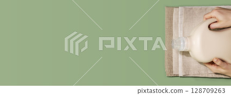 Eco friendly washing banner. Female hands hold bottle with washing gel or fabric softener on a textile napkins on a green background with copy space. Laundry day. Flat lay, banner size 128709263