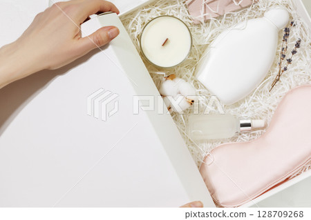 Female hands hold a white gift box with sleep care set. Preparing self care package, craft gift box with mask for sleep, aroma candle and bubble bath. Relaxing at home and take care concept. Closeup 128709268