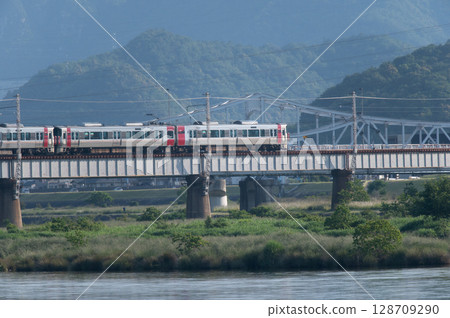 [Hiroshima Kabe Line] JR 227 Series train (Red Wing) running on a railway bridge with green mountains in the background 128709290