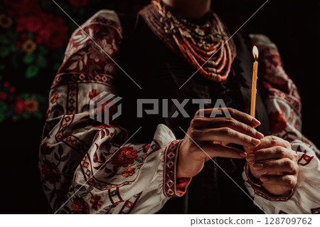 Ukrainian woman in traditional clothing holding candle in dark room interior 128709762
