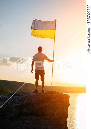 Man holding Ukrainian flag on cliff at sunset with scenic landscape view Man holding Ukrainian flag on cliff at sunset with scenic landscape view 128709765