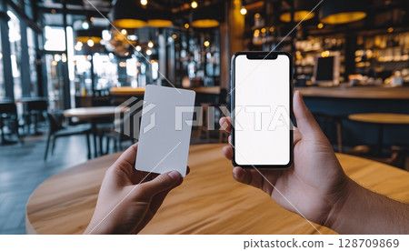 Person Holding Blank Card and Smartphone with Empty Screen in Modern Cafe Setting Person Holding Blank Card and Smartphone with Empty Screen in Modern Cafe Setting 128709869