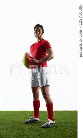Confident female rugby player posing with ball on green field background 128710586