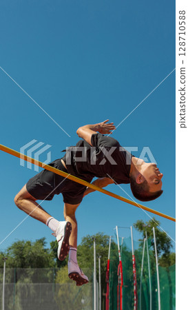 Athlete executing high jump beneath clear blue sky at outdoor track area 128710588