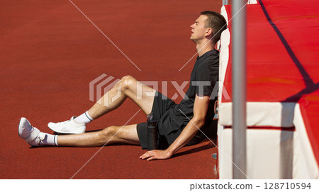 Athlete resting on track field under bright sunlight against high jump mat Athlete resting on track field under bright sunlight against high jump mat 128710594
