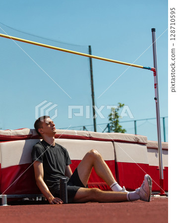 Track athlete resting against high jump mat under sunny blue sky 128710595