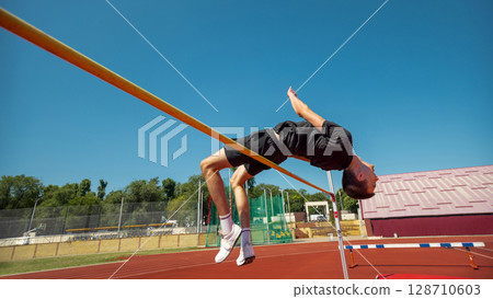 Athlete executing high jump on track beneath clear blue sky Athlete executing high jump on track beneath clear blue sky 128710603