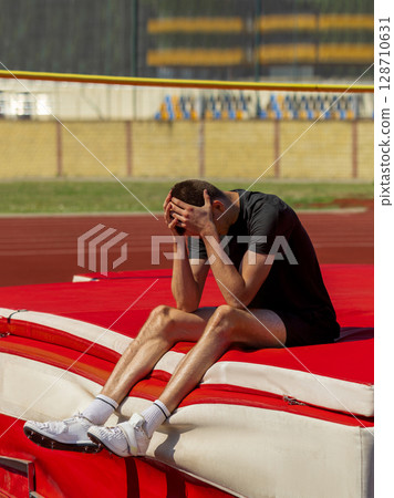 Exhausted athlete sitting on high jump mat during track and field practice 128710631
