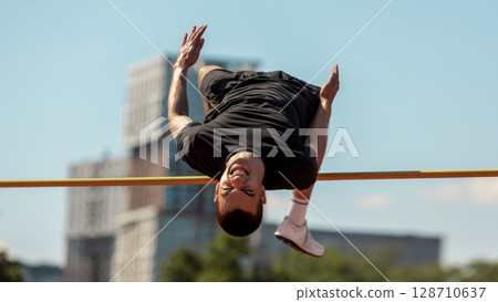 Athlete executes high jump with urban scenery on sunny day 128710637