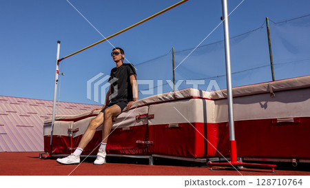 Athlete in sunglasses resting on high jump mat under clear blue sky at track field Athlete in sunglasses resting on high jump mat under clear blue sky at track field 128710764