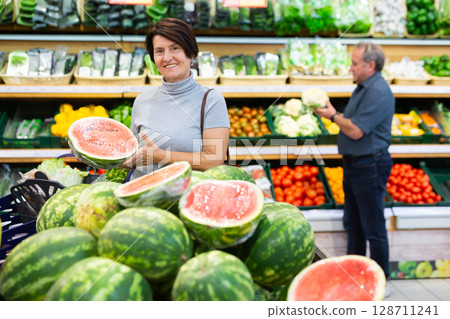 elderly woman chooses watermelon in fruit and vegetable section 128711241
