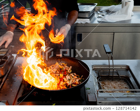 Chef cooking stir fry with flames outdoors in professional kitchen 128714142