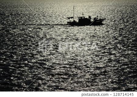 The silhouette of a fishing boat sailing on the sparkling sea in the setting sun 128714145
