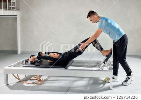 Woman practicing Pilates on a reformer with instructor in a bright studio Woman practicing Pilates on a reformer with instructor in a bright studio 128714226