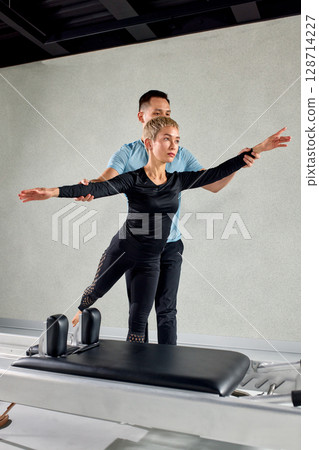 Professional fitness trainer guiding client during a pilates session on equipment Professional fitness trainer guiding client during a pilates session on equipment 128714227