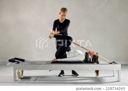 Professional Pilates trainer assists a client with reformer stretching exercise 128714243