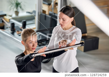 Fitness trainer assisting a client with equipment during a gym workout Fitness trainer assisting a client with equipment during a gym workout 128714259