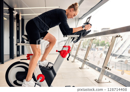 Woman exercising on a stationary bike indoors with a city view. 128714408
