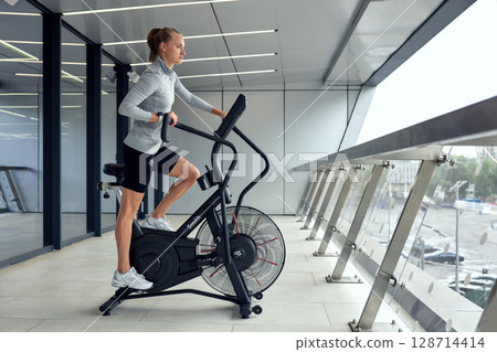 Woman exercising on a modern air bike in a sleek gym interior Woman exercising on a modern air bike in a sleek gym interior 128714414