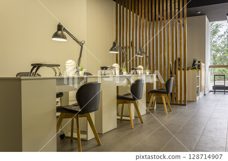 Interior of a modern beauty salon, nail area. Manicure workplace with lamps and cozy chairs in the barbershop. Concept of interior design for hairdressers and nail artists. Nail salon Interior of a modern beauty salon, nail area. Manicure workplace with lamps and cozy chairs in the barbershop. Concept of interior design for hairdressers and nail artists. Nail salon 128714907