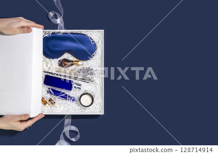 Men holiday healthy gift box. Female hands close a gift box with sleep care set for men. Preparing self care package with mask for sleep, candle, aroma sticks, oil and cosmetics. Flat lay, Copy space 128714914