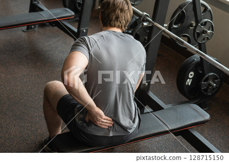 Athlete or young man sitting with pain in lower back at gym with muscle tension. Arm, inflammation or exercise with discomfort at fitness center. Close up. Athlete or young man sitting with pain in lower back at gym with muscle tension. Arm, inflammation or exercise with discomfort at fitness center. Close up. 128715150