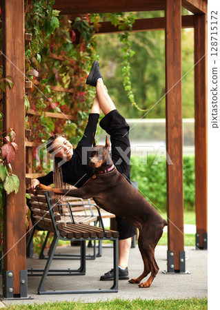 Woman practicing yoga outdoors with a Doberman in a park setting 128715172