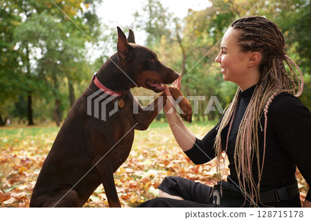 Woman Communicating with Loyal Doberman in a Sunlit Leafy Park Setting 128715178