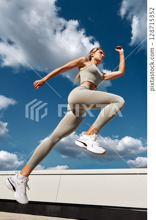Woman in sportswear leaping outside with a blue sky and clouds background 128715352