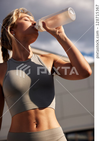 Athletic woman drinking from water bottle after outdoor workout session 128715367