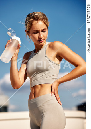 Fitness woman hydrating with water bottle during outdoor workout under sunny skies Fitness woman hydrating with water bottle during outdoor workout under sunny skies 128715376