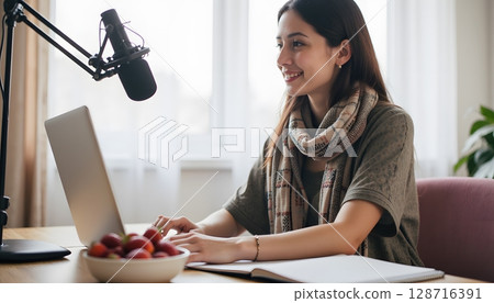 Young woman working on laptop at home office with microphone and fresh fruits on table 128716391